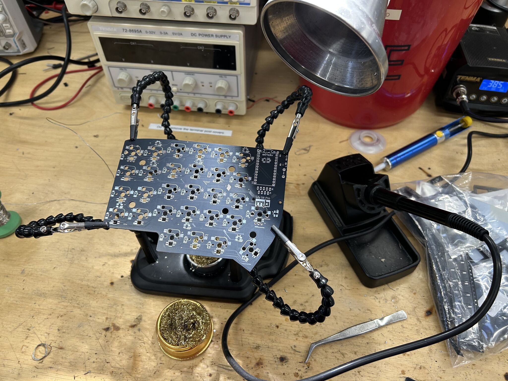 The PCB held in a third-hand vice on the workbench, mid-assembly. Helping hands, sponge, soldering iron, and a bench power supply for current-limited bring-up.